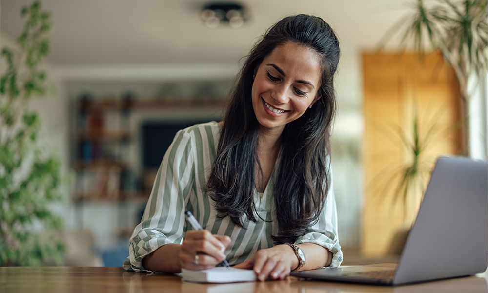 Eine lächelnde junge Frau arbeitet an Ihrem Computer und macht sich Notizen.